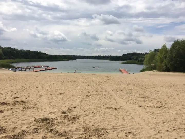 Piaskowa plaża nad jeziorem, z pomostami i łódkami. W oddali las, chmury na niebie. Idealne miejsce na wypoczynek nad wodą.