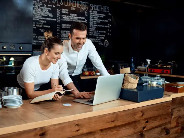 Pareja revisa menú en laptop, planificando su negocio de hostelería en España.