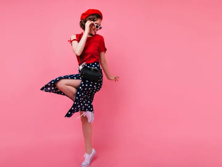 Kobieta w stylu pin-up w czerwonym berecie i sukience w grochy skacze na różowym tle.