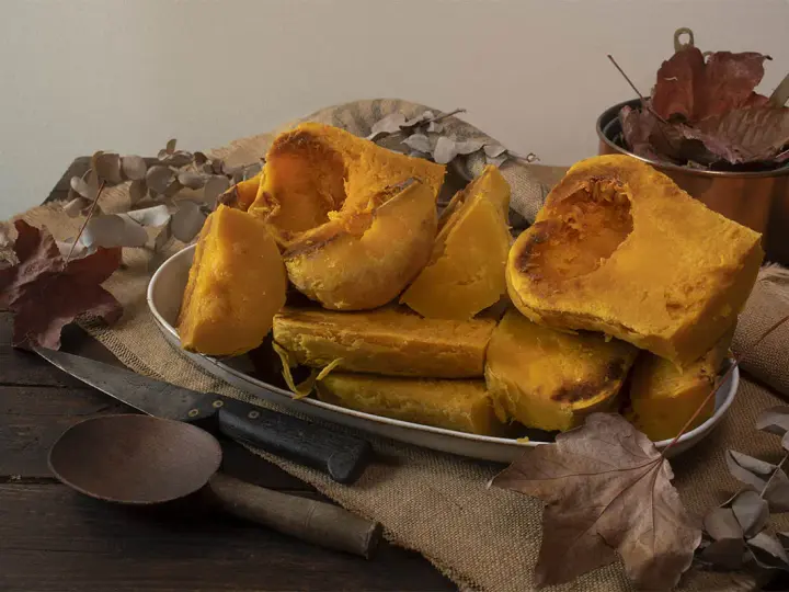 Trozos de calabaza asada, tierna y dorada, servidos en un plato blanco sobre arpillera. Acompañada de utensilios rústicos y hojas secas, evoca el sabor del otoño.