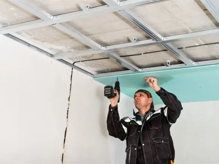 Un obrero instala un **pladur techo** con una taladradora. La estructura metálica ya está fijada al techo original.