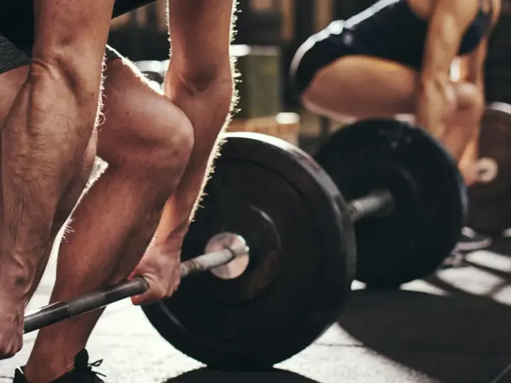 Personer lyfter skivstång i gym. Frågan "hur lång tid tar det att bygga muskler" besvaras med konsekvent träning.