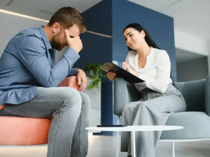 Mężczyzna w stresie podczas sesji, która może być częścią terapii uzależnień online. Terapeutaka notuje.