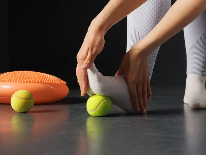 Manos masajeando pie con pelota de tenis, parte de ejercicios fascitis plantar. A su lado, un disco de equilibrio naranja.