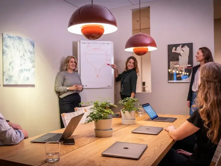 Ett team diskuterar strategier vid ett konferensbord med flera laptops. En kvinna pekar på en trattmodell på en whiteboard. Ups datorn är en del av deras effektiva arbetsflöde.