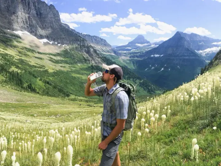 Randonneur buvant de l'eau au milieu d'un champ de fleurs blanches, avec des montagnes majestueuses en arrière-plan. Il espère que cela soulagera sa crampe cuisse.