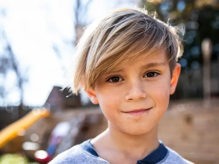 Ein Junge mit blonden Haaren, die nach vorne fallen, lächelt in die Kamera.