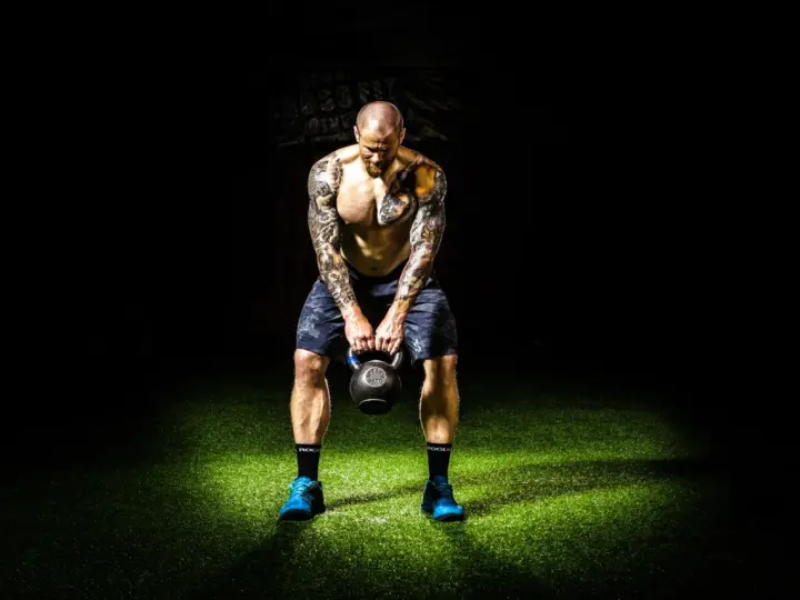 Hombre tatuado realiza press militar con kettlebell, enfocado en su entrenamiento.