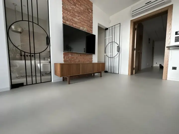 Nowoczesne wnętrze z betonową podłogą, co to jest mikrocement, która nadaje mu surowego charakteru. W tle widać łazienkę i salon.