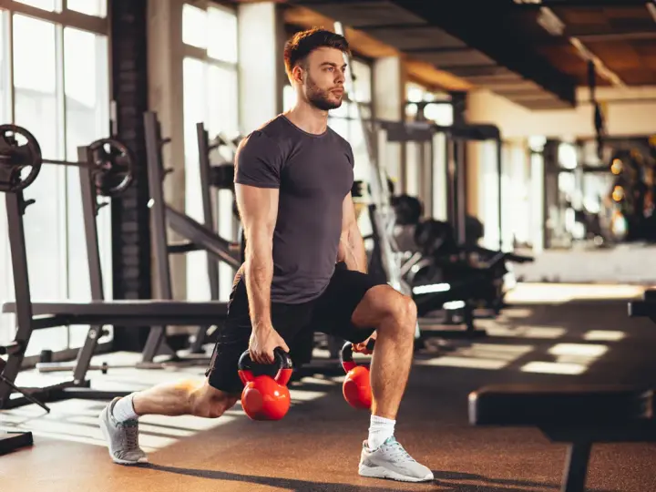 Mężczyzna wykonuje wykroki z kettlebellami, realizując swój plan treningowy na siłownię.