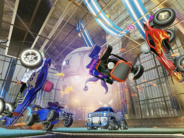 Trzy samochody z gry Rocket League w powietrzu, jeden czerwony, jeden niebieski i jeden pomarańczowy, na tle hali.