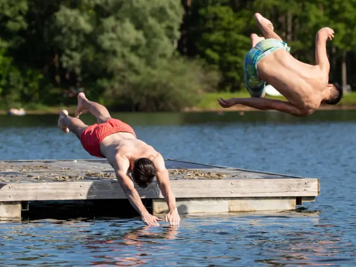 Zwei Männer springen vom Steg in den Sieglarer See. Einer macht einen Salto, der andere taucht gerade ins Wasser.