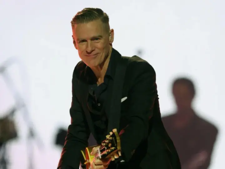 Bryan Adams w czarnym garniturze gra na gitarze. Sprawdź jego nadchodzące wydarzenia i koncerty.
