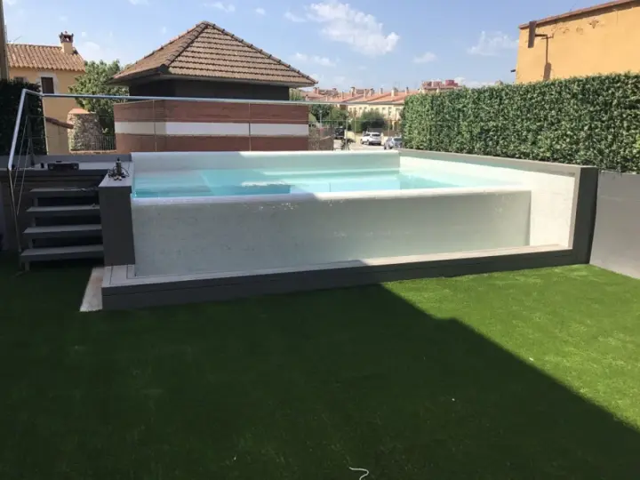 Moderna piscina con mosaico blanco, rodeada de césped artificial y setos. Ideal para decoración de terrazas con piscina.