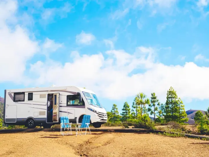 Un camper bianco è parcheggiato in un'area sterrata con due sedie blu e un tavolino. Sullo sfondo, pini e colline sotto un cielo azzurro con nuvole bianche. Un luogo ideale per un soggiorno in stile piscin'e janas.