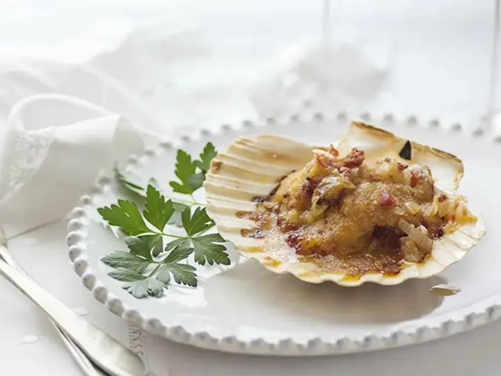 Vieiras gratinadas con tocino y perejil, servidas en su concha sobre un plato blanco.