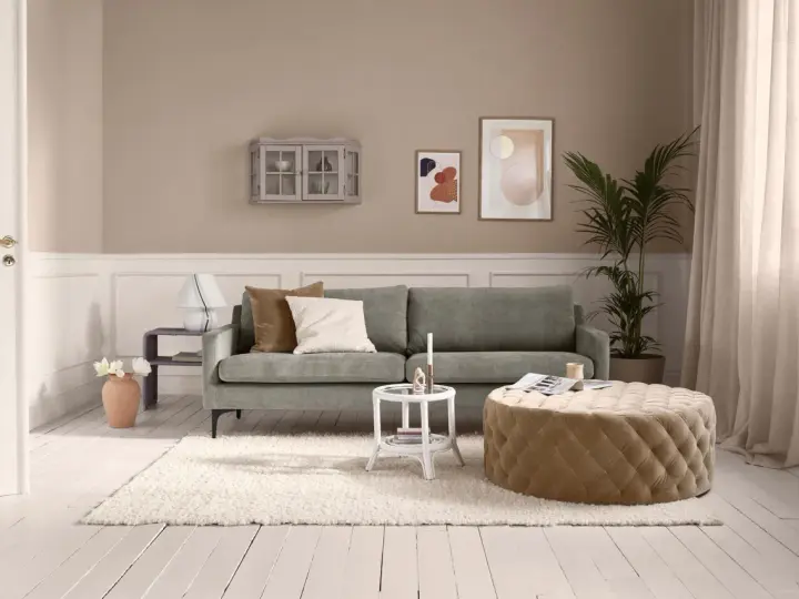 Ein Wohnzimmer mit salbeigrünem Sofa. Braune und cremefarbene Kissen, ein beiger Hocker und eine helle Wand zeigen, welche Farbe passt zu Salbei.