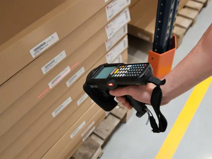 Mano escaneando cajas apiladas con un dispositivo, usando la **fórmula de Wilson** para optimizar el inventario.