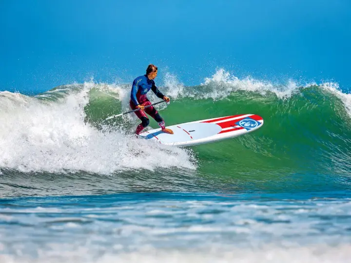 Ein Mann im Neoprenanzug surft auf einem SUP-Board eine grüne Welle. Das SUP-Wellenreiten erfordert Balance und Geschick.