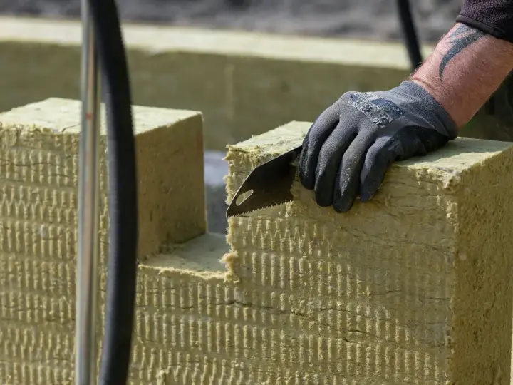 Ręka w rękawicy tnie piłą wełnę skalną 10. Materiał izolacyjny jest cięty na budowie.