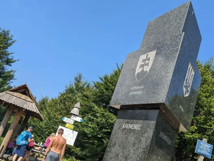 Trójstyk granic Bieszczady: monumentalny obelisk z herbami Słowacji i Ukrainy, obok drewniana wiata i turyści.
