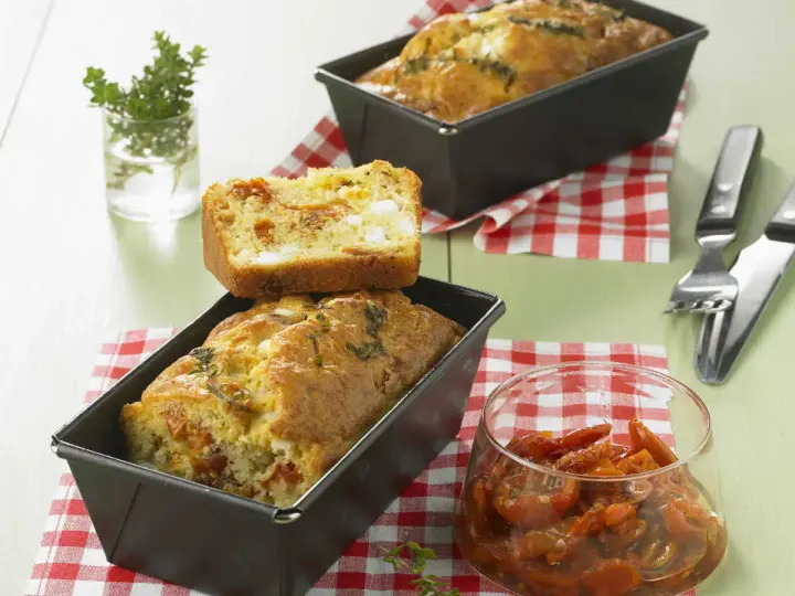 Cake tomates séchées feta, un délice salé aux herbes fraîches. Une tranche est posée sur un cake encore dans son moule.