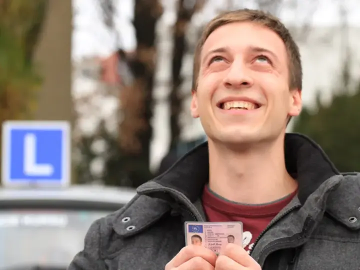 Szczęśliwy młody mężczyzna trzyma prawo jazdy kategorii B, z niebieskim znakiem "L" na dachu samochodu w tle.
