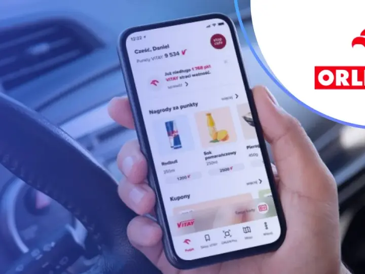 Dłoń trzyma smartfon z aplikacją ORLEN Pay, pokazującą punkty VITAY i nagrody. Kierownica samochodu w tle.