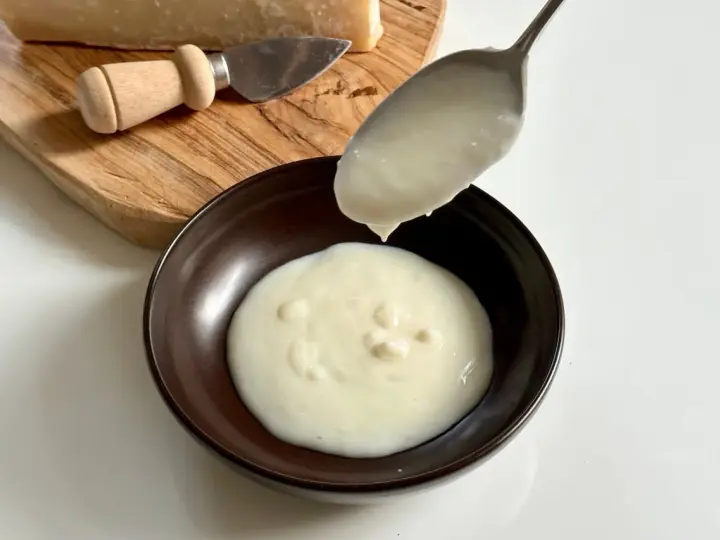 Una cucchiaiata di cremosa fonduta di parmigiano cade in una ciotola. Sul tagliere, un pezzo di Parmigiano Reggiano e un coltellino.