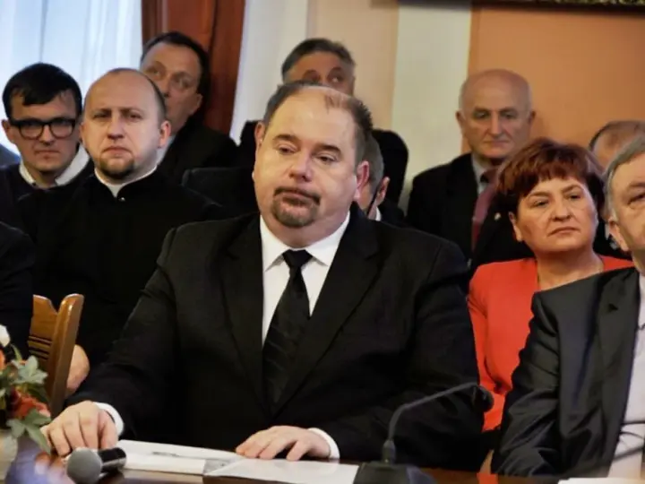 Jarosław Paluch w czarnym garniturze siedzi przy stole, otoczony innymi osobami.