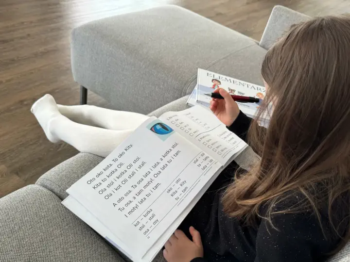 Dziewczynka z długimi włosami uczy się czytać z elementarza. Na stronach widać proste teksty, idealne do nauki czytania dla 7 latka.