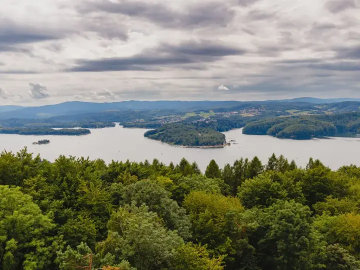 Widok na jezioro otoczone lasami, z górami w tle. Na pierwszym planie bujna zieleń drzew, a dalej spokojna tafla wody i zabudowania. Nad wszystkim chmury, jakby biała góra na niebie.