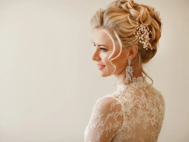 Eleganckie uczesania na wesele średnie włosy: blondynka w koronkowej sukni z perłową ozdobą i długimi kolczykami.
