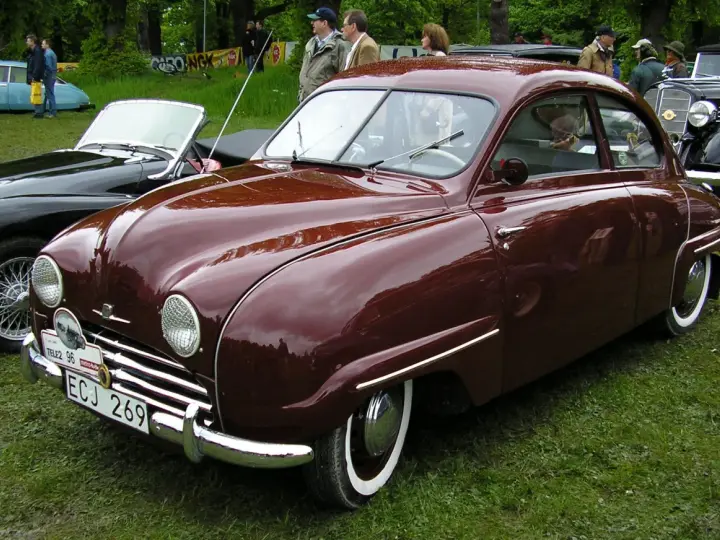 En vinröd Saab 92 står på gräs, med en svart cabriolet bredvid. Bilen har en klassisk design och en svensk registreringsskylt.