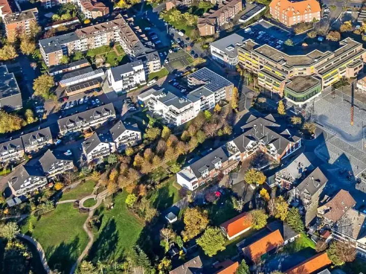 Luftaufnahme eines Wohngebiets mit Mehrfamilienhäusern und Einfamilienhäusern. Die Bäume zeigen Herbstlaub. Ein zentraler Platz ist zu sehen. Hier könnte man den Mietspiegel Voerde studieren.