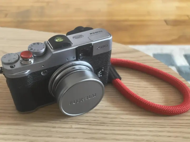 Srebrny aparat Fujifilm X20 z czerwoną smyczą leży na drewnianym blacie.