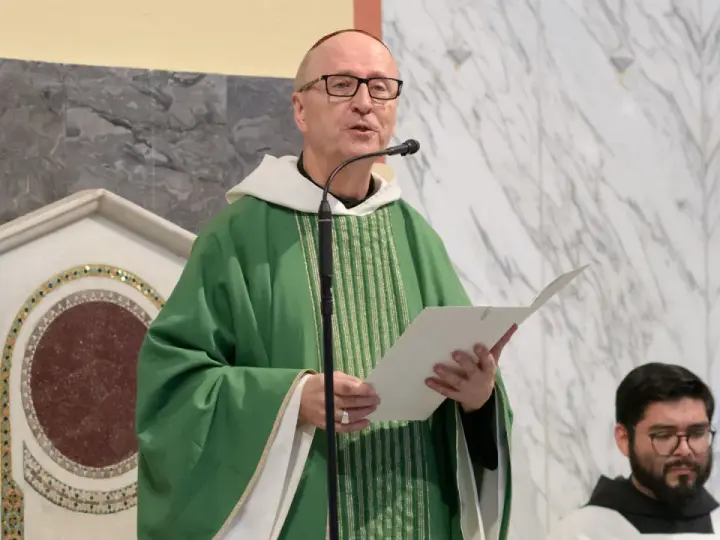 A man with a Benedictine title, wearing a green chasuble, speaks into a microphone while holding a paper.