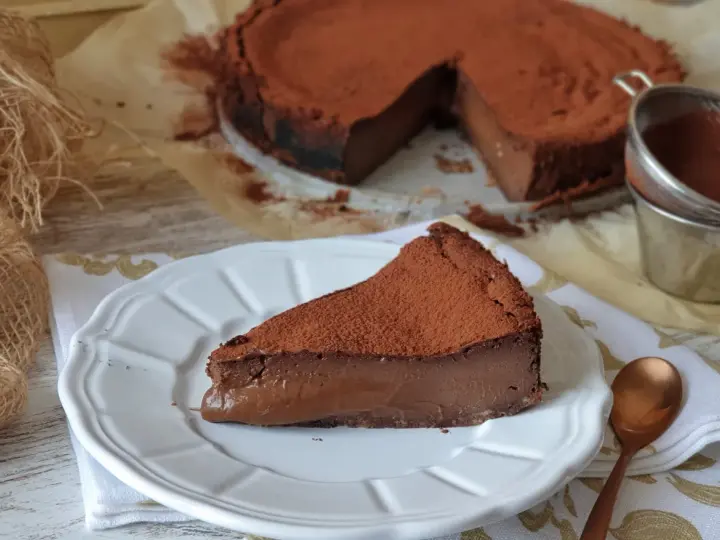 Una porción de tarta de chocolate, cubierta de cacao en polvo, reposa en un plato blanco. El resto de la tarta, también espolvoreada, se ve al fondo.
