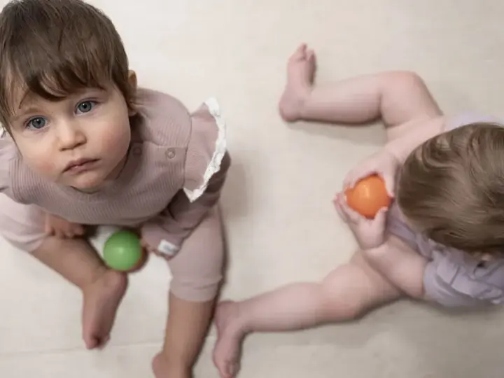 Dwa niemowlęta, jedno 9 miesięczne dziecko z niebieskimi oczami, bawi się zieloną piłeczką.