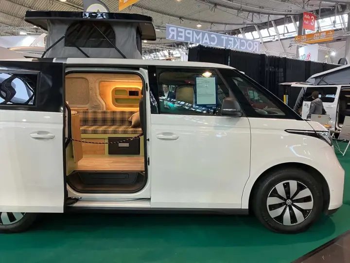Vit ID Buzz camper van med uppfällt tak och öppen skjutdörr som visar en mysig interiör med rutig soffa och trädetaljer.