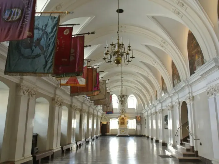 Długi korytarz z łukami i żyrandolami, ozdobiony historycznymi sztandarami. To wnętrze klasztoru na Jasnej Górze, gdzie każdy baner opowiada historię zakonu paulinów.