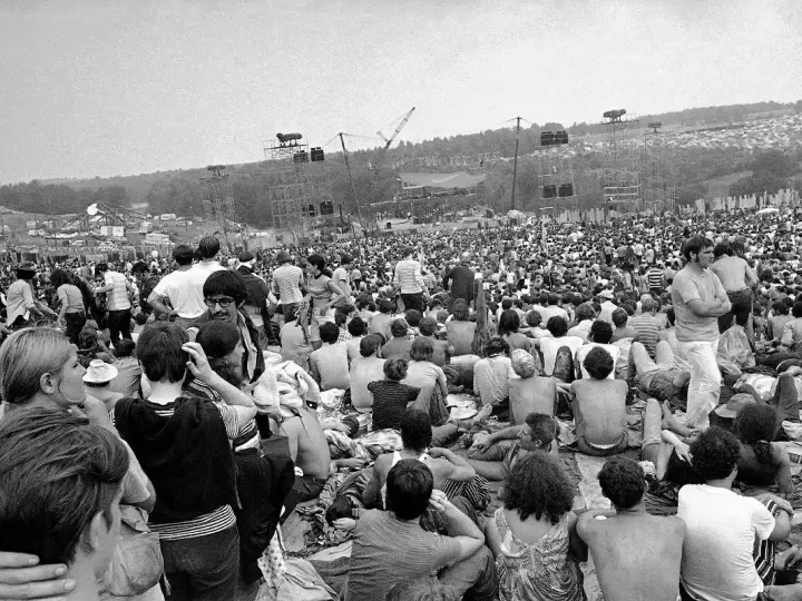 Tysiące ludzi siedzi na trawie, patrząc na scenę festiwalu. To jest Woodstock, gdzie jest muzyka i wolność.