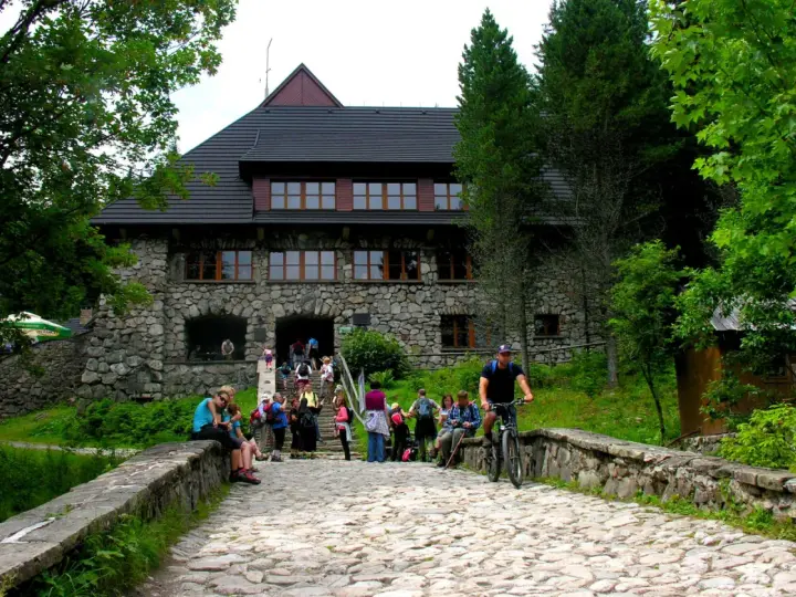 Kamienny budynek schroniska w Dolinie Chochołowskiej z ludźmi na schodach i rowerzystą na kamiennej drodze.