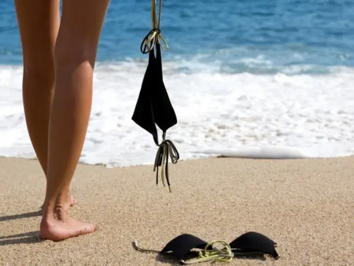Nogi na piaszczystej plaży, obok czarny kostium kąpielowy. Fale obmywają brzeg. Idealne miejsce na naturystyczną plażę.