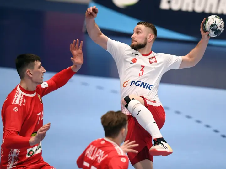 Gracz w piłkę ręczną w skoku, z piłką w dłoni, celuje do bramki. Jego przeciwnik próbuje go zatrzymać.