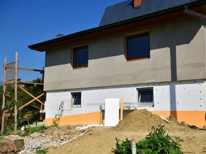 Budynek w trakcie ocieplania styropianem. Widoczne okna, rusztowanie i ziemia. Trzeba uważać na **mostek termiczny** przy oknach.