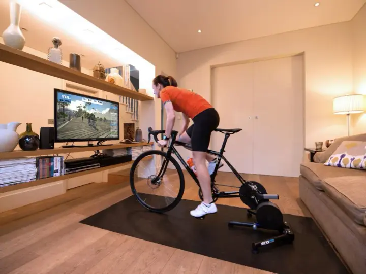Woman in her home cycling pain cave, training indoors on a bike trainer while watching a virtual race on a large screen.