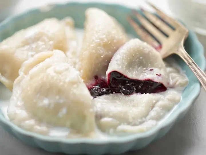 Pyszne pierogi z jagodami, polane śmietaną, w niebieskiej miseczce. Widoczny widelec.