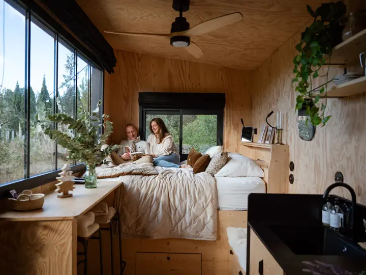 Un couple lit dans un lit douillet, profitant de l'aménagement ingénieux d'une tiny house avec vue sur la nature.