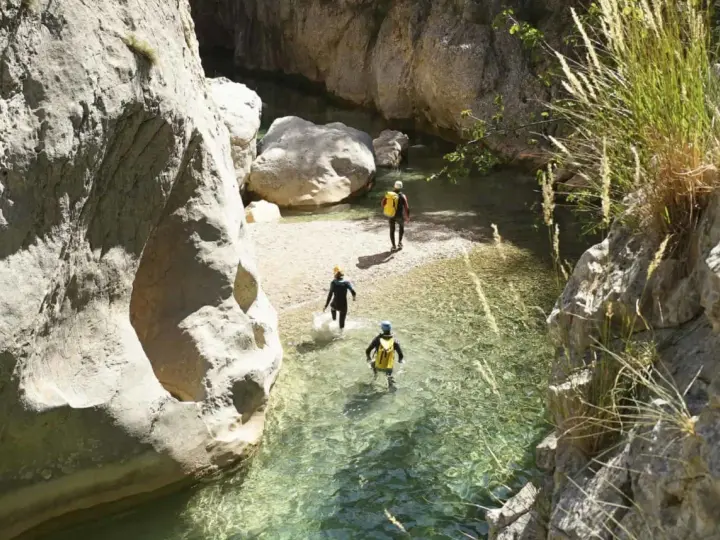 Trois personnes en combinaison font du canyoning dans une rivière aux eaux claires, entourées de falaises rocheuses.